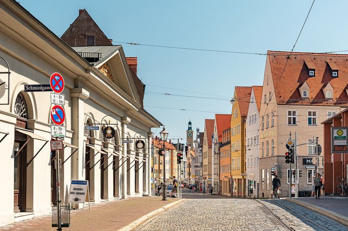 A cobbled old-town street in Augsburg lined with colourful stepped-gable houses, tram wires overhead and a church tower closing the view at the far end.