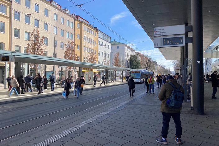 The Königsplatz interchange in Augsburg: tram tracks set into the paving, stop canopies and a departure board, commuters crossing and a city bus waiting under a clear sky.