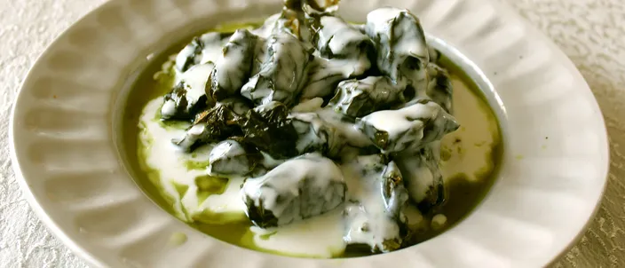 A white fluted plate of small, tightly rolled vine-leaf dolma parcels in olive oil, topped with a spoonful of thick white yogurt.