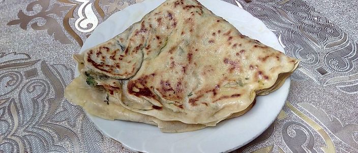 A folded qutab flatbread on a white plate, browned and glistening with butter, its green herb filling visible at the seam.