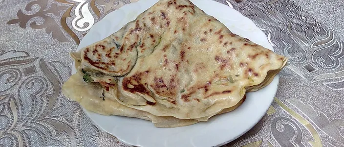 A folded qutab flatbread on a white plate, browned and glistening with butter, its green herb filling visible at the seam.