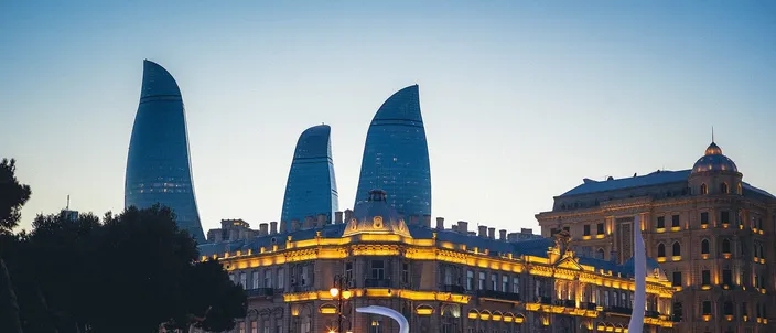 The three Flame Towers illuminated at dusk rising behind an ornate stone building on Baku Boulevard, with three white swan-shaped fountain sculptures in the foreground pool.