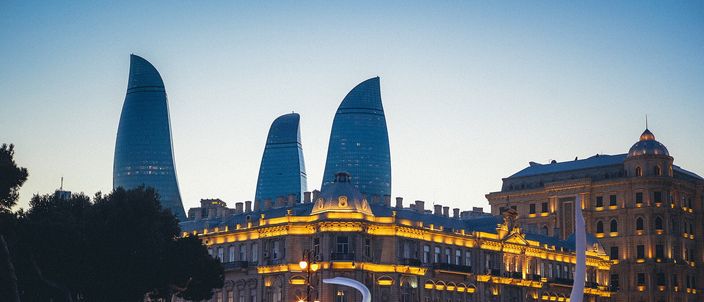 The three Flame Towers illuminated at dusk rising behind an ornate stone building on Baku Boulevard, with three white swan-shaped fountain sculptures in the foreground pool.