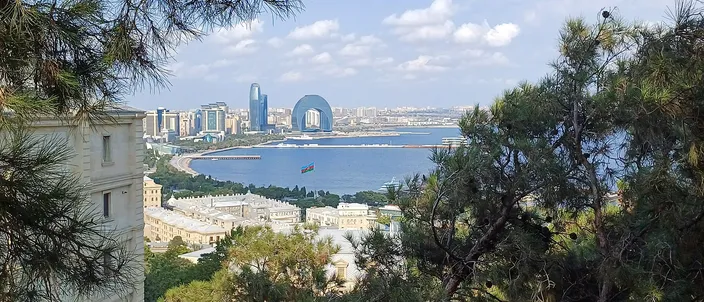An elevated view of Baku's bay, seaside boulevard and skyline through pine branches, the crescent-shaped waterfront building at center