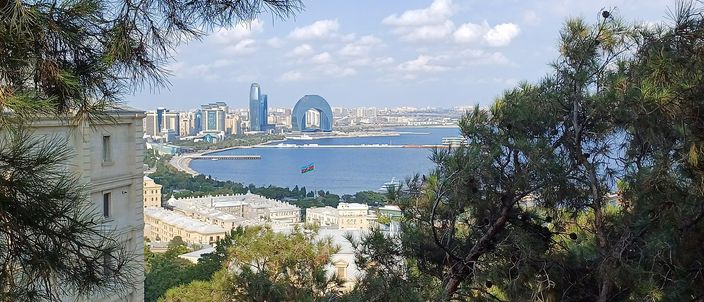 An elevated view of Baku's bay, seaside boulevard and skyline through pine branches, the crescent-shaped waterfront building at centre