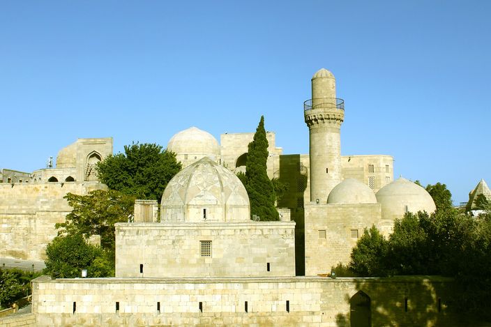 The Palace of the Shirvanshahs complex — pale-stone domed pavilions, courtyards and a slender minaret — seen from below against a clear sky.