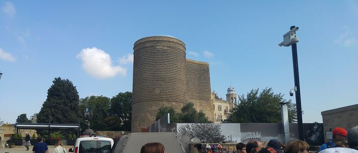 The Maiden Tower's cylindrical stone drum and narrower buttress rising above the Old City square, with visitors gathered at its base under a clear sky.