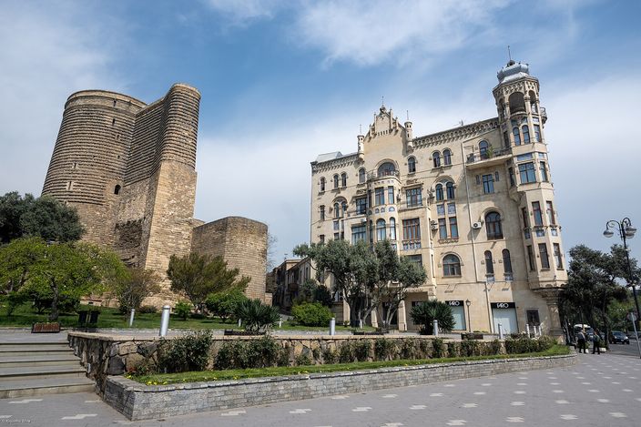 The cylindrical stone Maiden Tower beside an ornate Art Nouveau mansion inside Baku's walled Old City, seen across a park path under a clear blue sky.