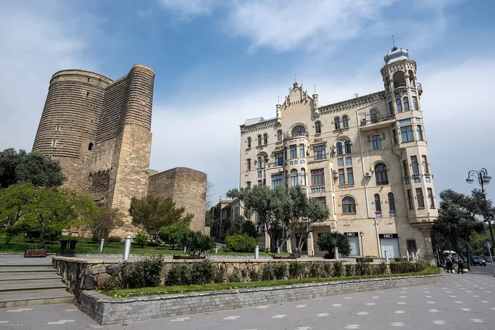 The cylindrical stone Maiden Tower beside an ornate Art Nouveau mansion inside Baku's walled Old City, seen across a park path under a clear blue sky.