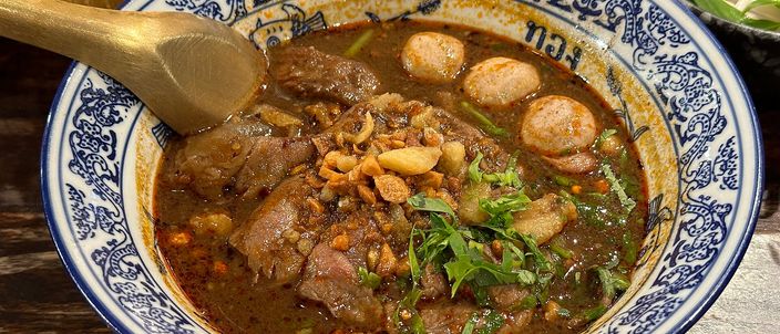 A blue-and-white patterned Thai bowl of boat noodles: dark, nearly black broth with sliced beef, a beef ball, braised shank and tendon, thin rice noodles, fried garlic and coriander, a brass spoon resting in it and a side plate of raw greens behind.