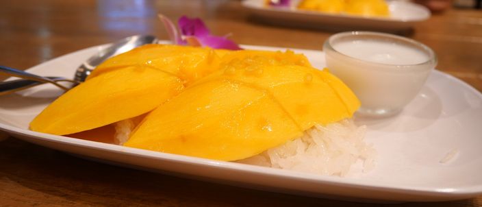 A white oval plate of mango sticky rice: two fans of ripe deep-yellow mango slices laid over a mound of coconut sticky rice, with a small cup of coconut cream and an orchid garnish beside it on a wooden table.