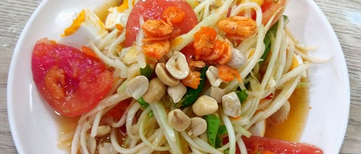 A white plate of som tam seen from above: shredded green papaya tossed with wedges of tomato, roasted peanuts, dried shrimp and pieces of salted egg in a glistening lime-and-fish-sauce dressing.