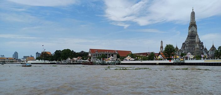 The Chao Phraya River in daylight with the tall white-and-gold prang of Wat Arun rising on the far bank beneath a blue sky with wispy clouds, boats on the churning brown water.