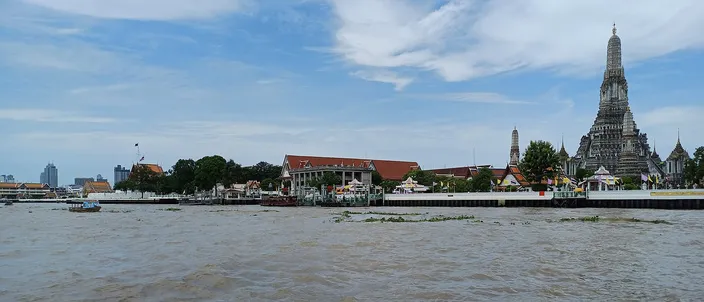 The Chao Phraya River in daylight with the tall white-and-gold prang of Wat Arun rising on the far bank beneath a blue sky with wispy clouds, boats on the churning brown water.
