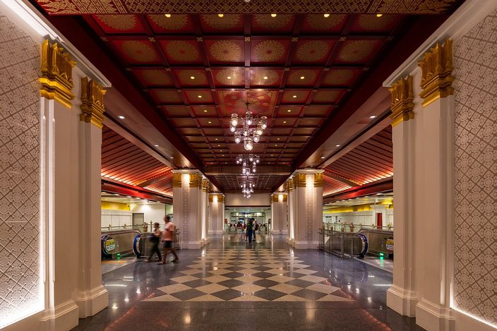 The underground concourse of Sanam Chai metro station in Bangkok: gilded columns with lotus capitals, a deep red coffered ceiling picked out in gold, hanging chandeliers, a black-and-white chequered floor and a few travellers walking towards the escalators.