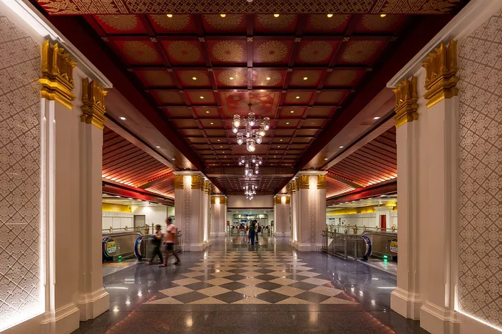 The underground concourse of Sanam Chai metro station in Bangkok: gilded columns with lotus capitals, a deep red coffered ceiling picked out in gold, hanging chandeliers, a black-and-white chequered floor and a few travelers walking towards the escalators.