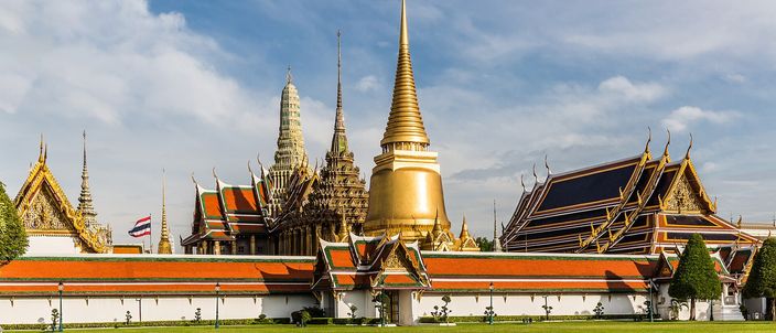 The outer court of the Grand Palace in Bangkok: a golden bell-shaped chedi flanked by tiered spires and orange-and-green temple roofs, seen across a mown lawn under a blue sky with scattered cloud.