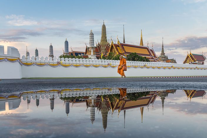 A monk in a saffron robe walking along the white outer wall of the Grand Palace in Bangkok in early light, the gilded chedi and tiered prangs of Wat Phra Kaew rising behind the wall, the whole scene mirrored in rainwater lying on the pavement.