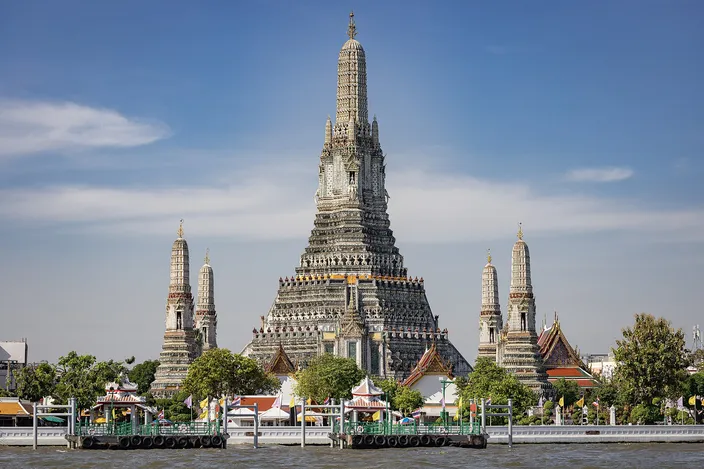 Wat Arun seen from the Chao Phraya: the tall porcelain-encrusted central prang with four smaller satellite prangs around it, pale gray against a blue sky, temple roofs and a green landing stage along the water in front.
