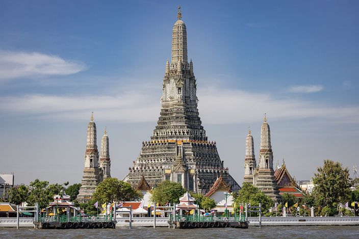 Wat Arun seen from the Chao Phraya: the tall porcelain-encrusted central prang with four smaller satellite prangs around it, pale grey against a blue sky, temple roofs and a green landing stage along the water in front.