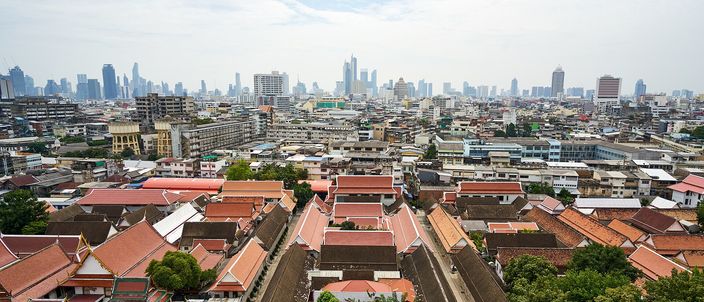 The old quarter of Bangkok seen from the terrace of the Golden Mount: red-tiled roofs and gilded temple gables among dense low-rise streets, running back to a distant line of high-rise towers under a pale overcast sky.