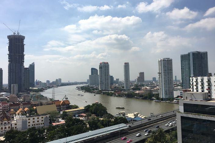 View over Bangkok from a Silom high-rise: the Chao Phraya River curving between clusters of high-rise towers under a bright cloudy sky, with an elevated roadway and traffic crossing the foreground.