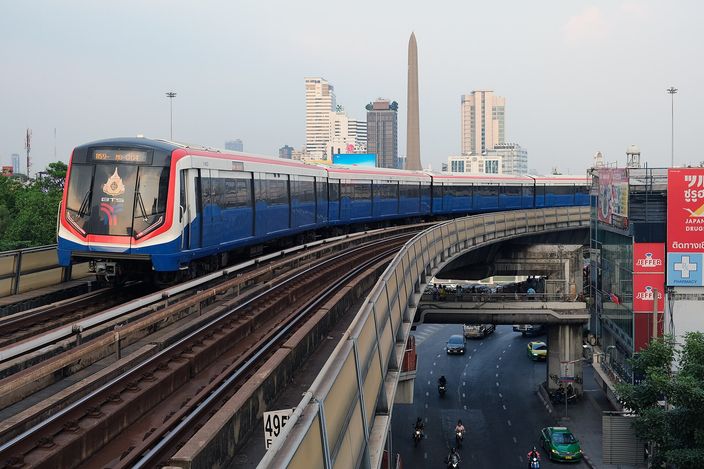 A red, white and blue BTS Skytrain train curving along an elevated concrete track above a multi-lane road with cars and motorbikes, city towers and the Victory Monument obelisk in the hazy background.