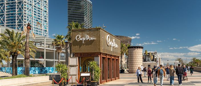A wood-shingled beach kiosk on Barcelona's Passeig Maritim de la Barceloneta, with palm trees, people walking the seafront promenade, and the sail-shaped W Hotel visible in the distance under a clear blue sky.