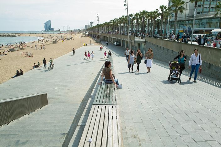 A view along Barcelona's seafront promenade: benches, palm trees and pedestrians on the right, and the wide pale-gold sand of the beach on the left scattered with sunbathers, with a tall sail-shaped hotel tower far down the coastline.