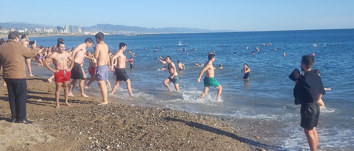 A crowd of people, some in short-sleeve wetsuits, run and wade into the sea on a bright winter morning; the low sun and hazy Barcelona coastline stretch away behind them.