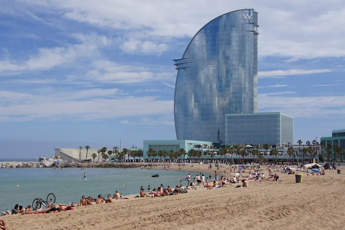 Sunbathers spread across a pale sandy beach in the foreground; the sea is calm and shallow near shore, and the sail-shaped glass tower of the W Hotel rises behind a low breakwater at the harbor end of the beach.
