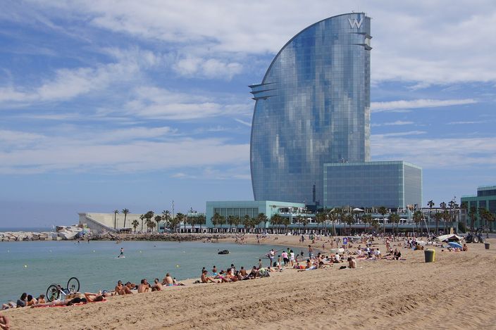 Sunbathers spread across a pale sandy beach in the foreground; the sea is calm and shallow near shore, and the sail-shaped glass tower of the W Hotel rises behind a low breakwater at the harbour end of the beach.