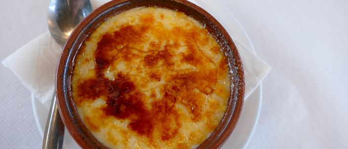 A traditional terracotta cazuela of crema catalana with a mottled, glossy burnt-sugar crust, a spoon resting beside it.