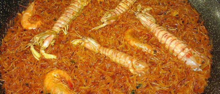 A wide, shallow pan of seafood fideuà: short toasted noodles in a reddish-golden broth topped with whole mantis shrimp and langoustines.