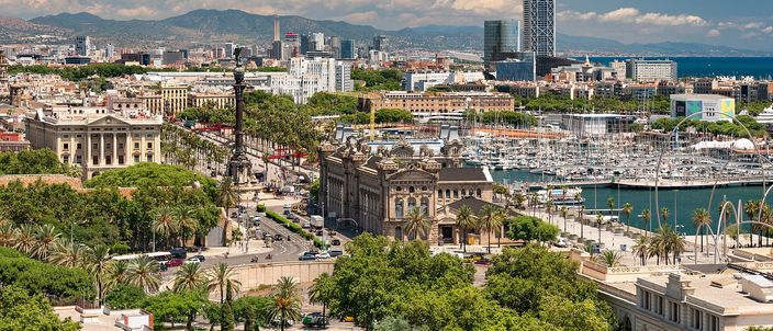 Barcelona seen from Montjuïc: the Columbus monument and neoclassical customs house among palm trees, the marina of Port Vell with moored yachts to the right, the sea and hills behind under a bright sky.