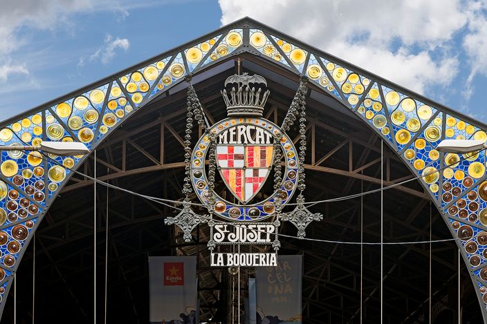 The modernista gable over the entrance to La Boqueria: stained-glass roundels in blue, amber and brown filling the ironwork of the arch, the crowned arms of Barcelona at the apex, and a wrought-iron sign beneath them reading Mercat St Josep - La Boqueria.