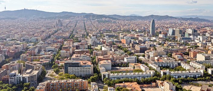 Barcelona from the air in warm afternoon light: the Eixample grid running inland in long straight avenues, the spires of the Sagrada Familia rising out of it left of centre, the bullet-shaped Torre Glories to the right, and the Collserola hills along the horizon.