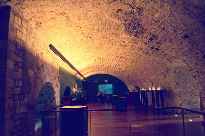 A long Romanesque barrel vault of rough stone over a hall of the former royal palace, lit warm from below, with a polished wooden floor, glass balustrades, low display cases along one side and a lit screen at the far end.