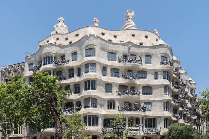 Casa Mila on its street corner: five floors of undulating pale cut stone wrapping the angle, wrought-iron balcony railings like tangled seaweed at every opening, and pale sculpted chimney stacks standing along the roofline against a clear blue sky.