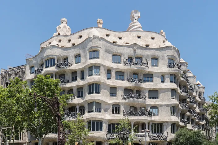 Casa Mila on its street corner: five floors of undulating pale cut stone wrapping the angle, wrought-iron balcony railings like tangled seaweed at every opening, and pale sculpted chimney stacks standing along the roofline against a clear blue sky.