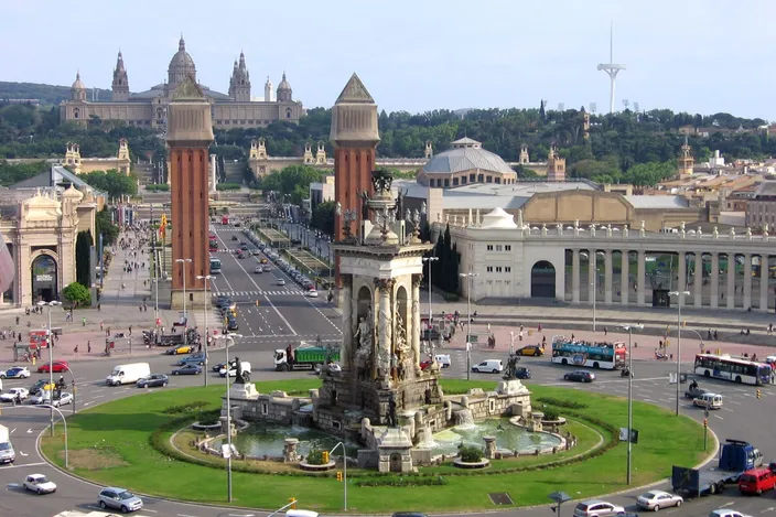 Placa d'Espanya seen from above: the 1929 fountain on its planted roundabout ringed with traffic, the two brick campaniles standing at the mouth of the avenue, and the avenue running up between the exhibition halls to the Palau Nacional on the hillside behind.