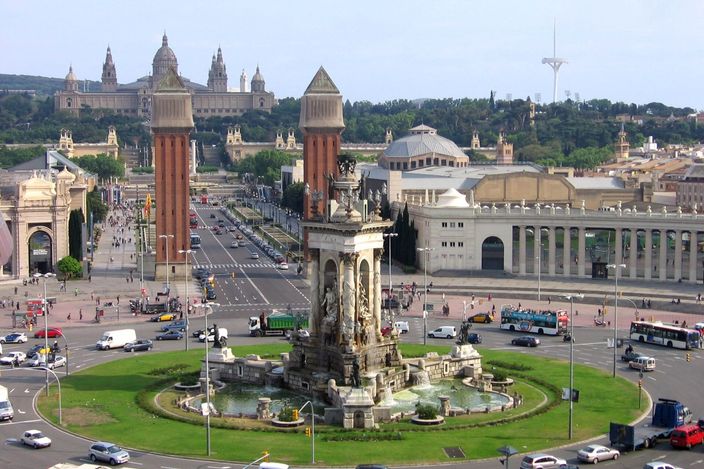 Placa d'Espanya seen from above: the 1929 fountain on its planted roundabout ringed with traffic, the two brick campaniles standing at the mouth of the avenue, and the avenue running up between the exhibition halls to the Palau Nacional on the hillside behind.