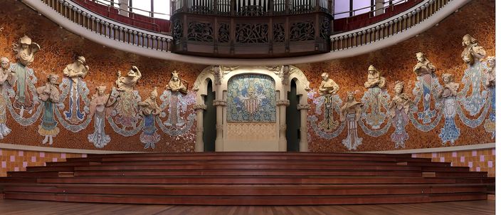 The stage wall of the Palau de la Musica Catalana: eighteen muses in relief and mosaic emerging from a wall of gold and terracotta tilework, a heraldic mosaic panel set between two arches at the centre, the pipe organ and a carved wooden gallery above them, and the empty wooden choir tiers in front.