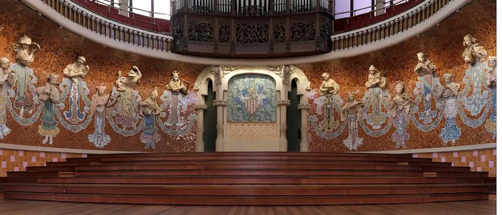 The stage wall of the Palau de la Musica Catalana: eighteen muses in relief and mosaic emerging from a wall of gold and terracotta tilework, a heraldic mosaic panel set between two arches at the center, the pipe organ and a carved wooden gallery above them, and the empty wooden choir tiers in front.