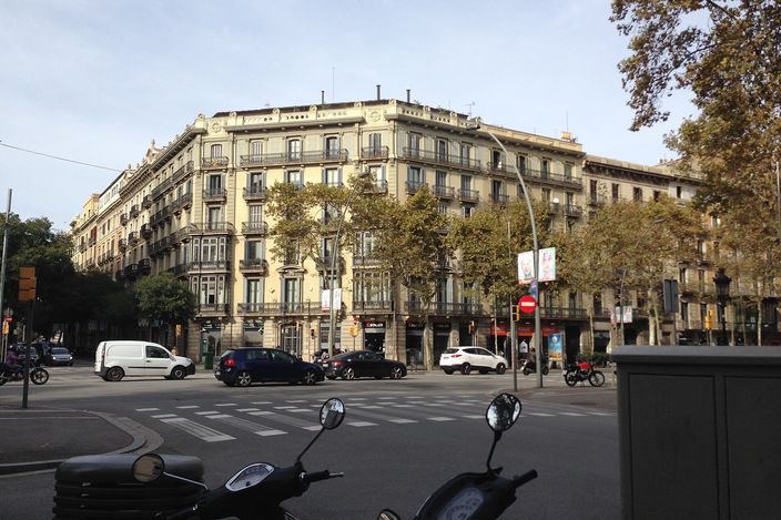 A corner block of ochre 19th-century Eixample buildings with wrought-iron balconies on the Gran Via de les Corts Catalanes, a wide street with a zebra crossing, cars and parked scooters in the foreground