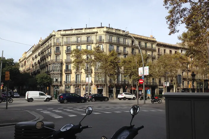 A corner block of ochre 19th-century Eixample buildings with wrought-iron balconies on the Gran Via de les Corts Catalanes, a wide street with a zebra crossing, cars and parked scooters in the foreground