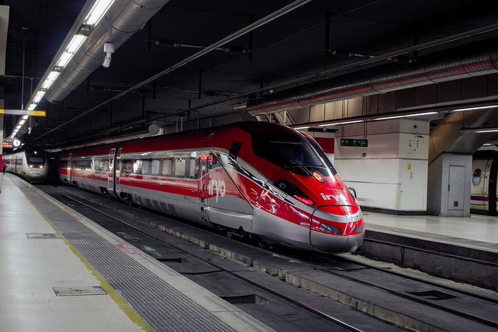 A red Iryo Frecciarossa high-speed train stands at a platform inside Barcelona Sants station, with another train visible on the adjacent track under the low station roof.