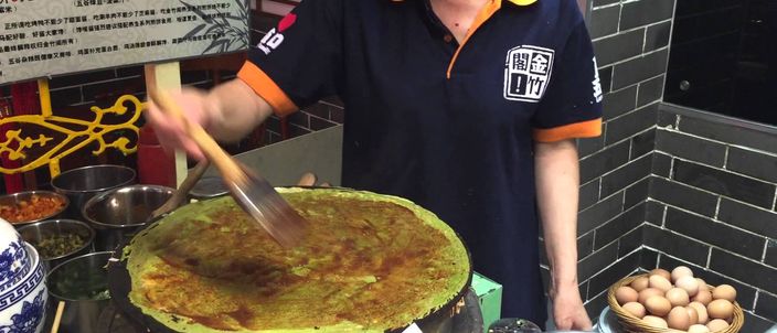 A street vendor spreading a thin jianbing crêpe on a round flat griddle, with egg smeared across the batter and dark sauce being brushed on, eggs and utensils to the side.