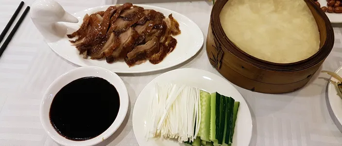 A Peking duck spread on a restaurant table: a plate of glossy sliced roast duck with crisp skin, a bamboo steamer of thin pancakes, a dish of dark sweet bean sauce, and batons of spring onion and cucumber.