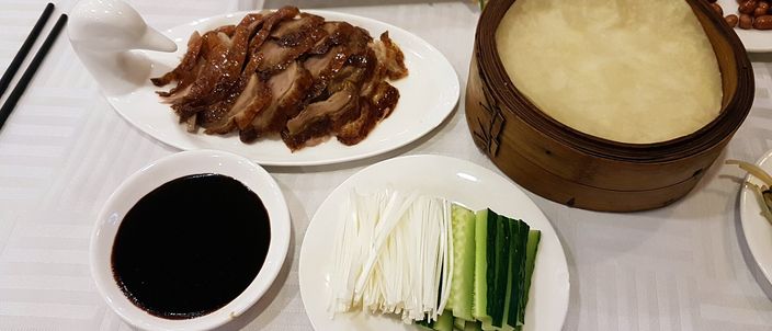 A Peking duck spread on a restaurant table: a plate of glossy sliced roast duck with crisp skin, a bamboo steamer of thin pancakes, a dish of dark sweet bean sauce, and batons of spring onion and cucumber.